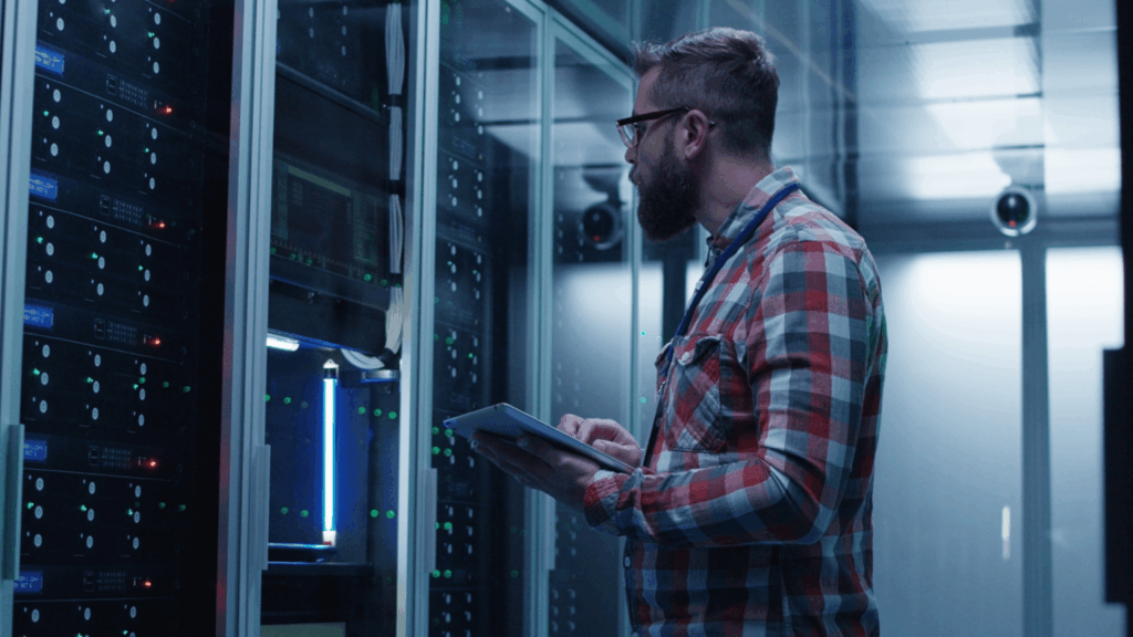 Homme vêtu d'une chemise à carreaux rouges et blancs, portant des lunettes, travaillant sur une tablette dans une salle serveur avec baies informatiques bleues